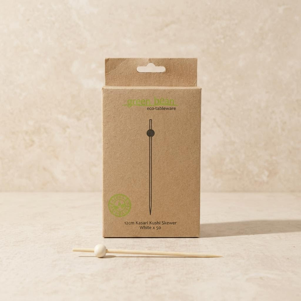 A kraft paper product box for a skewer set, with a single wooden skewer placed in front on a light stone surface. The box is tan with green and black printed text reading 'green bean eco-tableware' and '12cm kasar kubis skewer white x 50'.