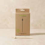A kraft paper product box for a skewer set, with a single wooden skewer placed in front on a light stone surface. The box is tan with green and black printed text reading 'green bean eco-tableware' and '12cm kasar kubis skewer white x 50'.