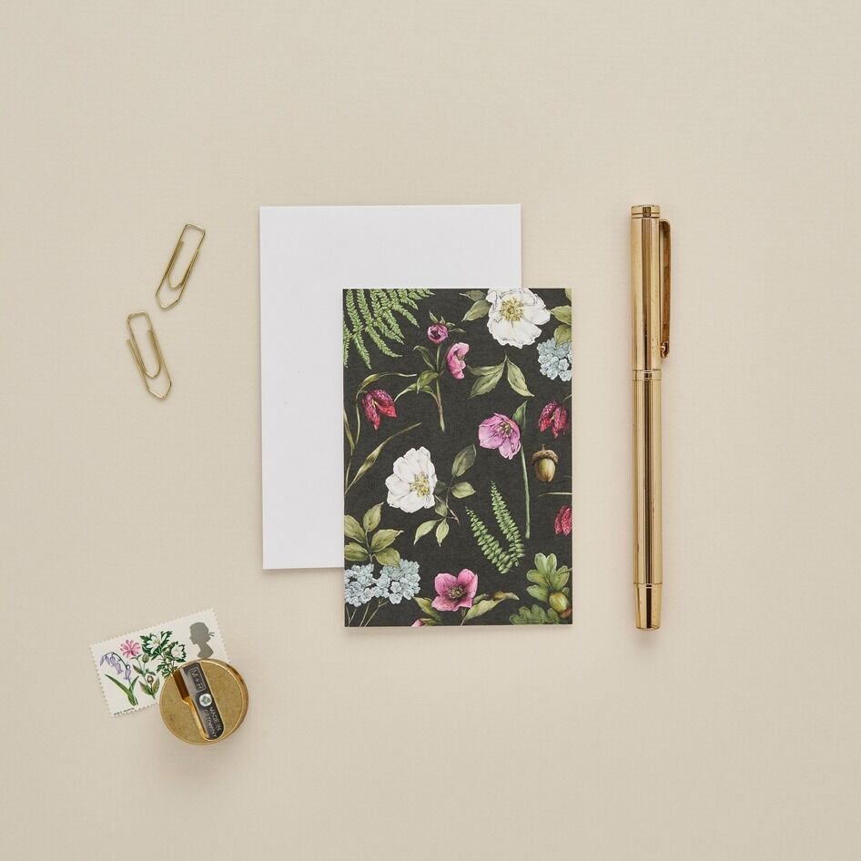 A floral patterned notepad or card with a dark background and pink, white, and green botanical print, styled on a beige surface with a white sheet, gold paperclips, a gold pen, and a small round pin.