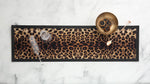 A rectangular leopard-print bar runner with black trim, styled on a white marble surface with a gold bowl, a cocktail spoon, a champagne flute, and a wine glass.