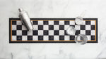 A rectangular bar mat with a black and white checkerboard pattern and a thin gold border, styled on a white marble surface with a cocktail shaker, strainer, and two glasses.