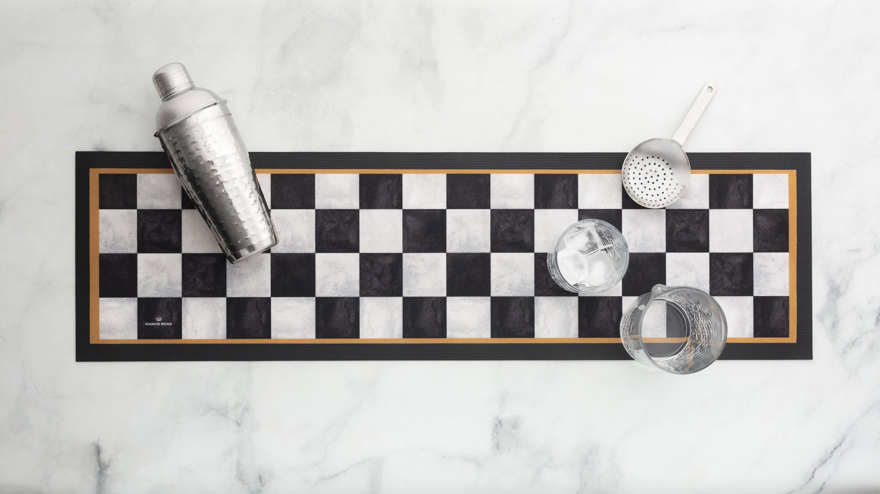 A rectangular bar mat with a black and white checkerboard pattern and a thin gold border, styled on a white marble surface with a cocktail shaker, strainer, and two glasses.