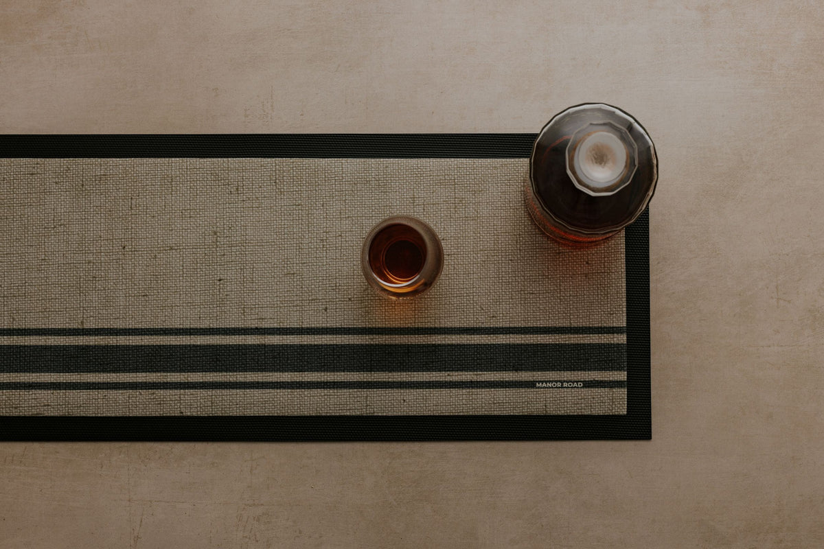 Rubber backed bar mat with linen woven look print. Glass bottle and drinking glass on the bar mat at a drinks station to catch spills. 