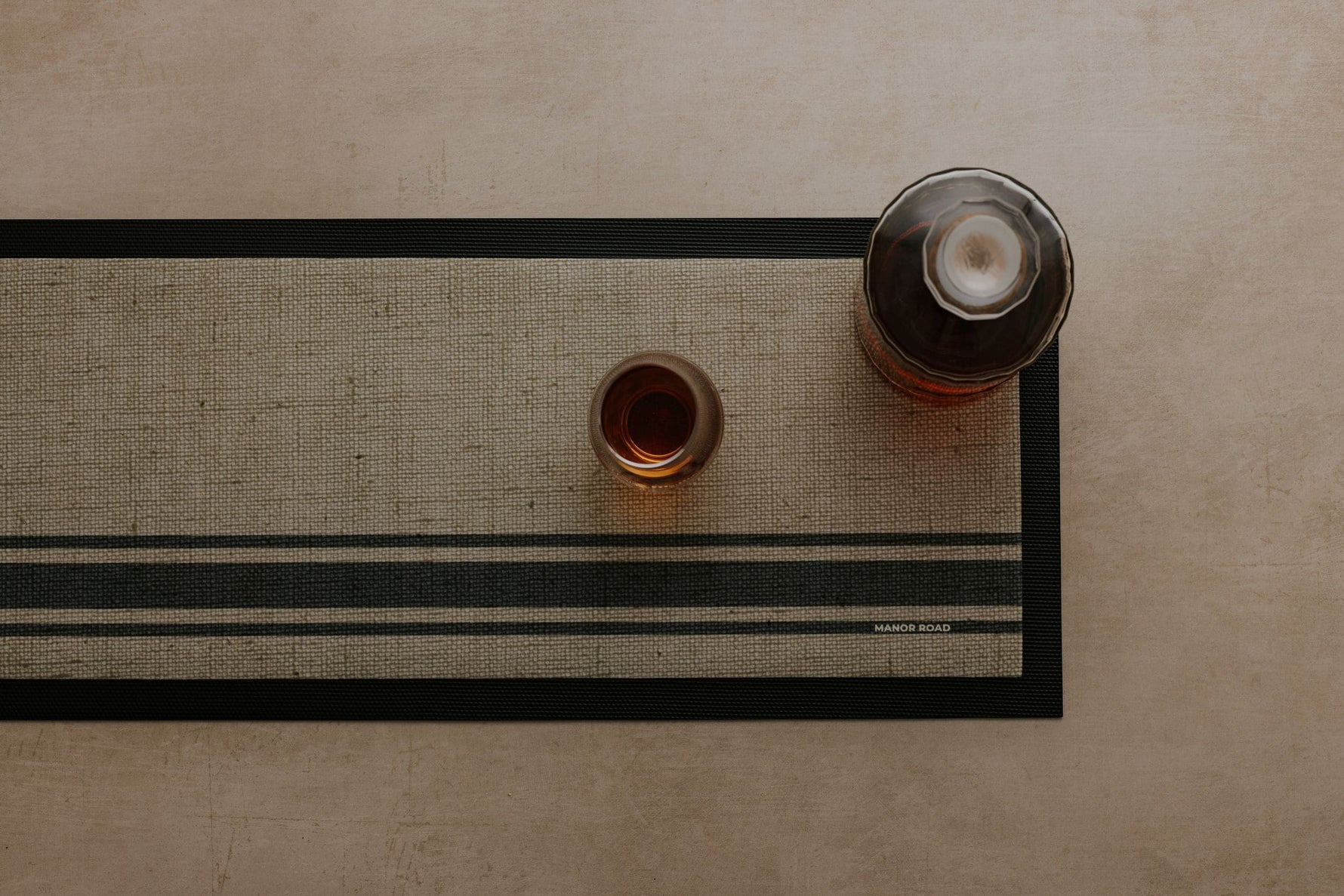 Rubber backed bar mat with linen woven look print. Glass bottle and drinking glass on the bar mat at a drinks station to catch spills. 