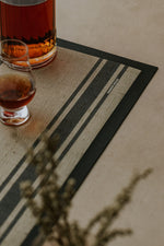 Two glasses of amber liquid on a striped rubber backed bar mat with a blurred plant in the foreground.