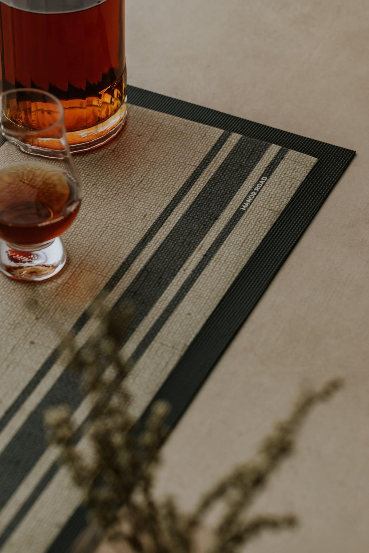 Two glasses of amber liquid on a striped rubber backed bar mat with a blurred plant in the foreground.