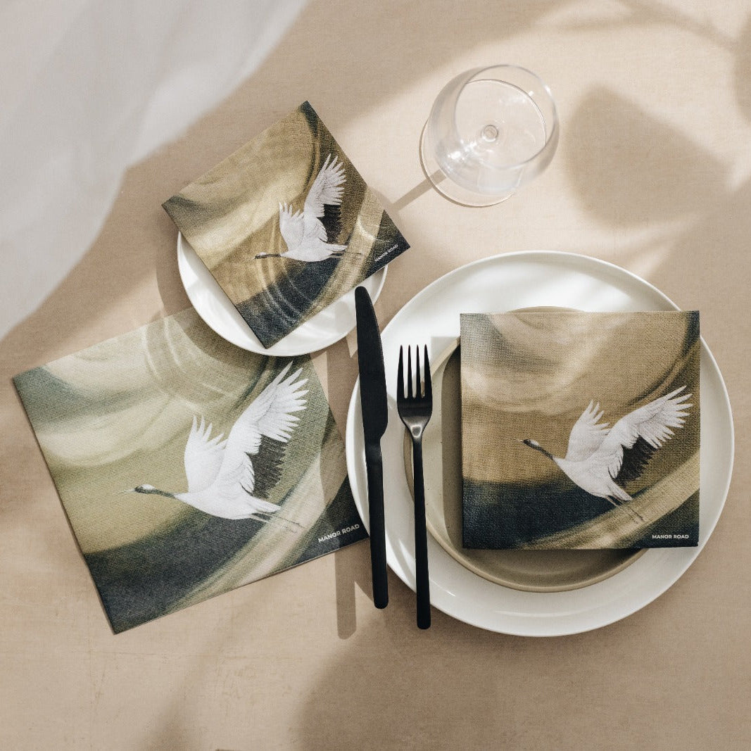 A styled flat-lay of a place setting featuring square napkins printed with a white flying crane on a beige and brown abstract background, arranged on white plates with black cutlery and a clear wine glass on a beige surface with soft shadows.