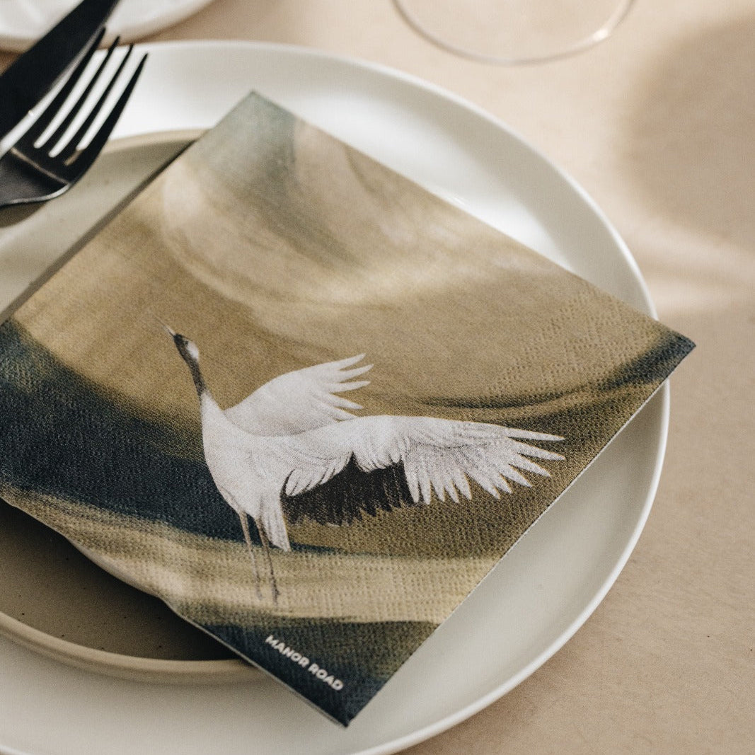 A folded paper napkin featuring a white crane over a muted landscape, styled on a white plate with a fork and knife on a beige table setting.