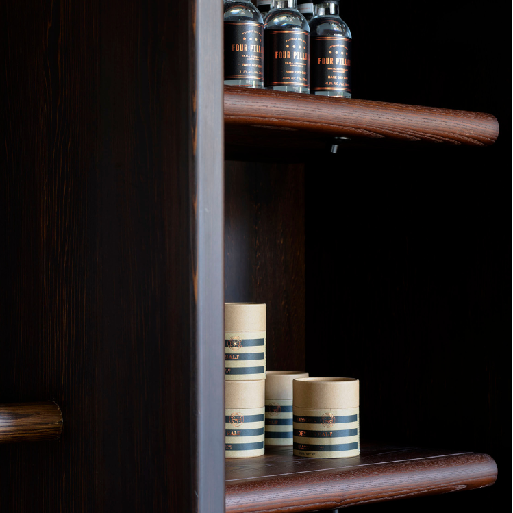 A close-up of a dark wood shelving unit with a matching horizontal handle, holding decorative bottles and stacked cylindrical containers on its shelves against a dark background.