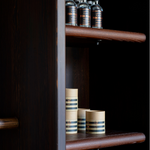 A close-up of a dark wood shelving unit with a matching horizontal handle, holding decorative bottles and stacked cylindrical containers on its shelves against a dark background.