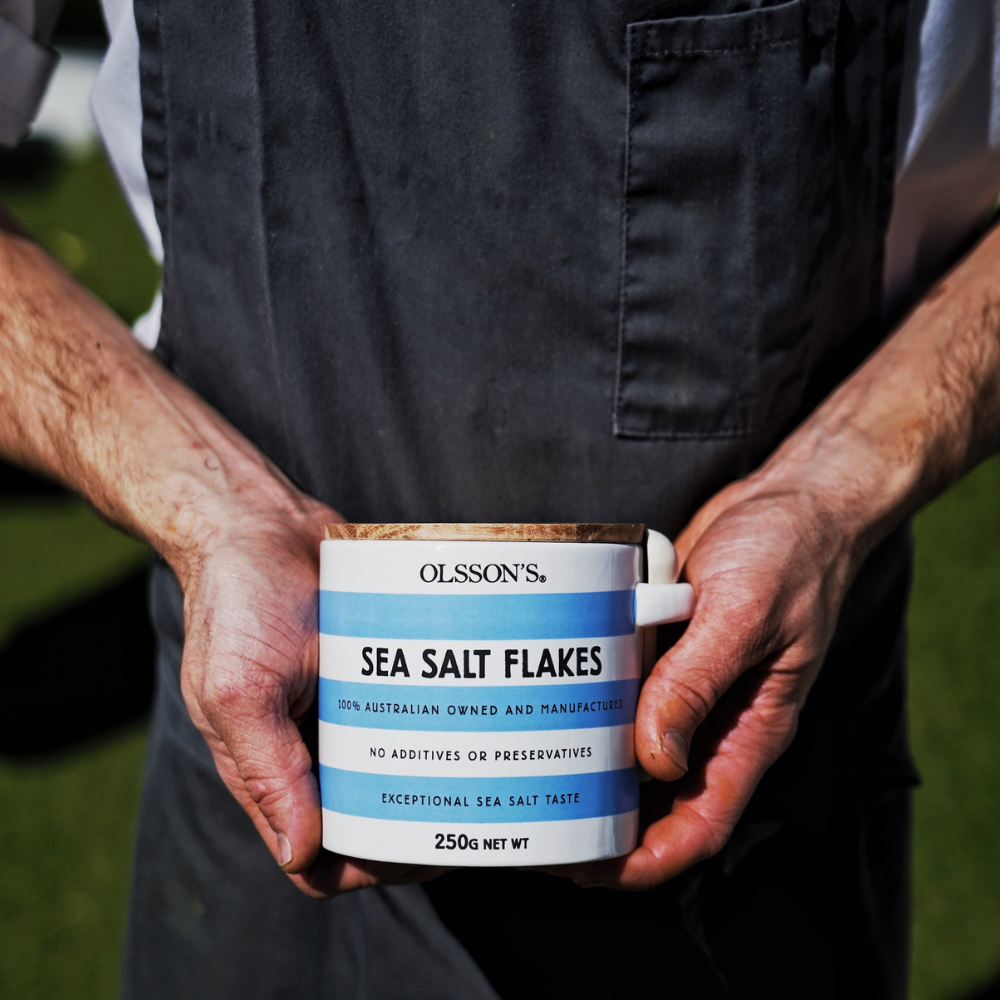 A person wearing a dark gray apron holds a white ceramic mug filled with Olsson's Sea Salt Flakes outdoors against a blurred green grass background.