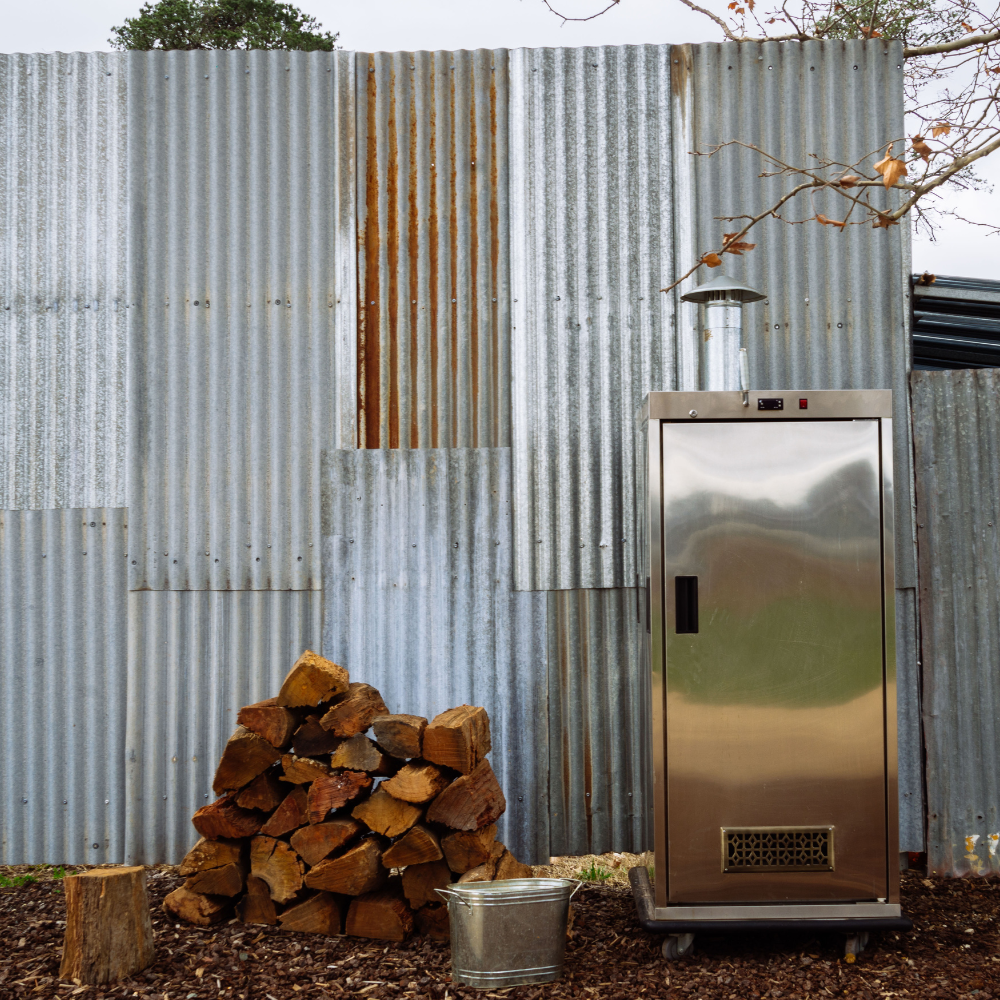 A stainless steel outdoor smoker or grill with a vent and chimney, set on a wood deck in front of a corrugated metal wall with stacked firewood and a metal bucket nearby.