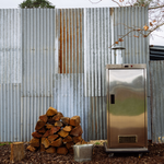 A stainless steel outdoor smoker or grill with a vent and chimney, set on a wood deck in front of a corrugated metal wall with stacked firewood and a metal bucket nearby.