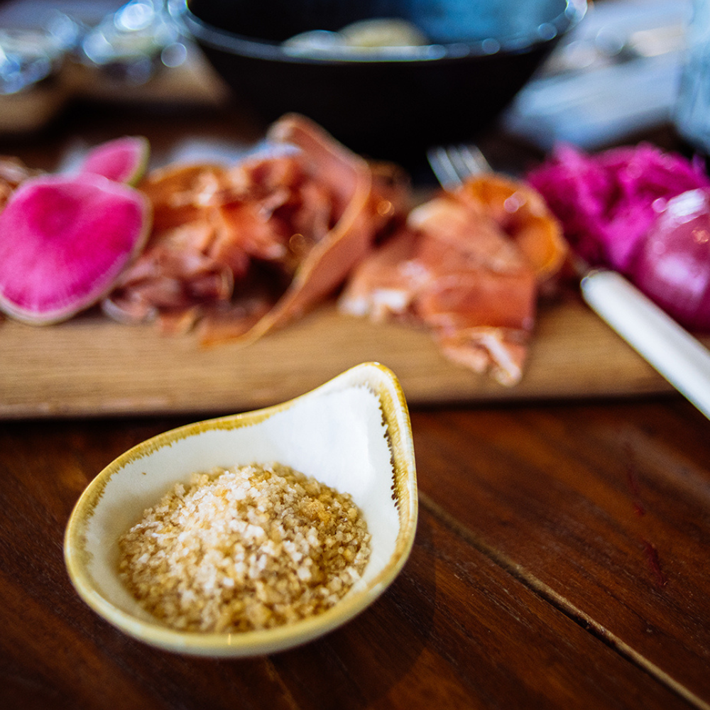 A small white ceramic bowl with a gold rim filled with coarse sea salt sits on a dark wooden table in the foreground, with a blurred board of sliced cured meat, pickled radishes, and a fork in the background.