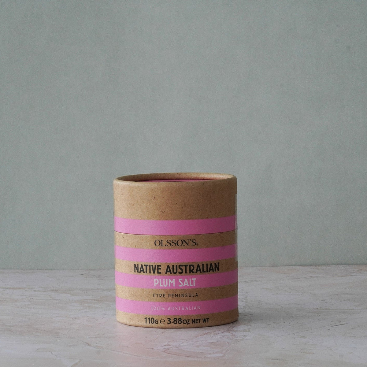 Olsson's Native Australian Ruby Salt in a kraft paper cylinder container with pink stripes, set on a light marble surface against a muted gray-green background.