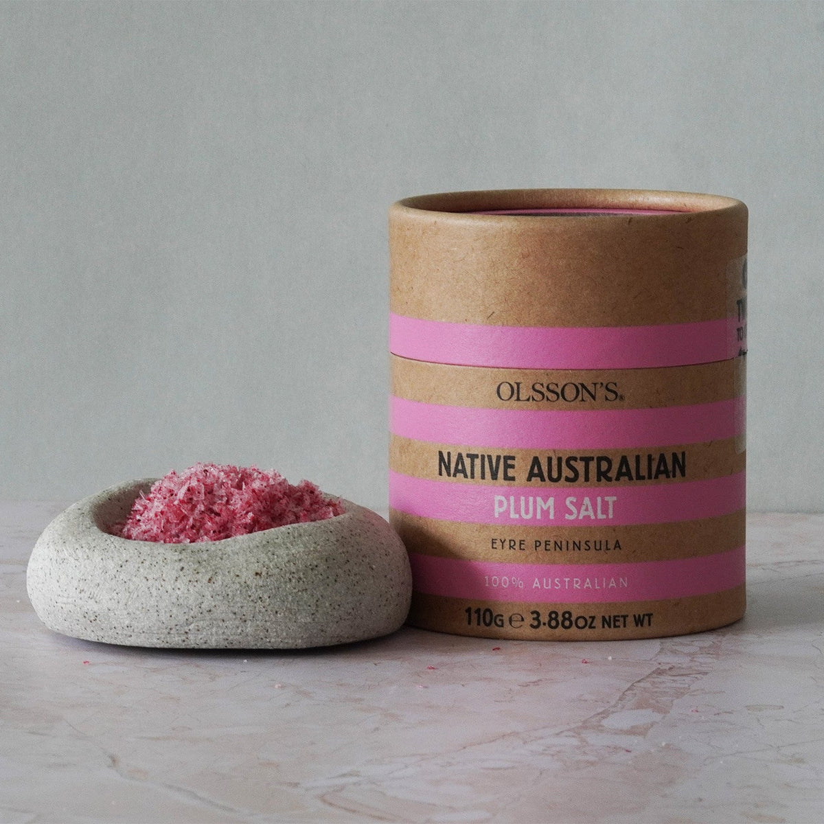 Olsson's Native Australian Plum Salt in a kraft paper cylinder with pink stripes, shown next to a small speckled stone dish filled with pink salt crystals on a light marble surface.
