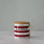 A ceramic salt container with a bamboo lid and a white wooden spoon, labeled 'Olsson's Red Gum Smoked Salt', set on a light marble surface against a neutral gray background.