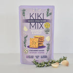 A package of Kiki Mix Rosemary & Garlic Seed Cracker in a light purple stand-up pouch, styled with garlic cloves and rosemary sprigs on a light gray background.