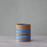 A cylindrical kraft paper container of Olsson's Sea Salt Flakes (Erik Peninsula) sits on a light stone surface against a textured gray wall.