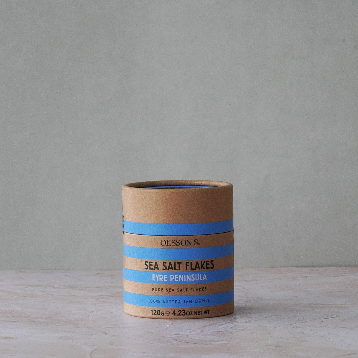 A cylindrical kraft paper container of Olsson's Sea Salt Flakes (Erik Peninsula) sits on a light stone surface against a textured gray wall.