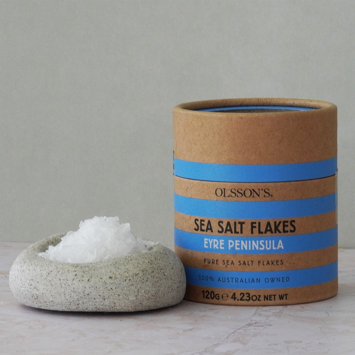 A cylindrical kraft paper container of Olsson's Fire Peninsula Sea Salt Flakes stands on a light stone surface next to a small speckled stone dish holding white salt flakes.