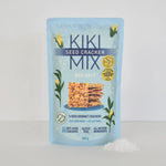A stand-up pouch of Kiki Seed Cracker Mix Sea Salt, 300g, standing on a white surface with a small pile of white sea salt in front. The packaging is light blue with dark blue text and illustrations of yellow flowers and green leaves.