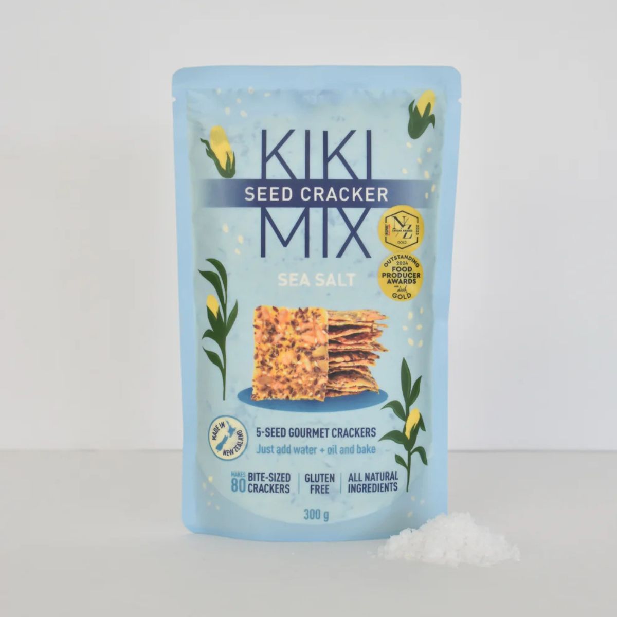 A stand-up pouch of Kiki Seed Cracker Mix Sea Salt, 300g, standing on a white surface with a small pile of white sea salt in front. The packaging is light blue with dark blue text and illustrations of yellow flowers and green leaves.