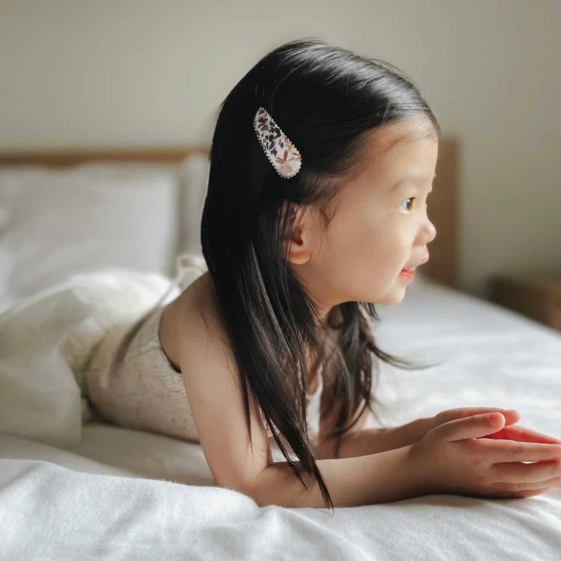 A young girl with long black hair lies on a white bed, wearing a decorative hair clip with a Liberty floral design in white, pink, and black.