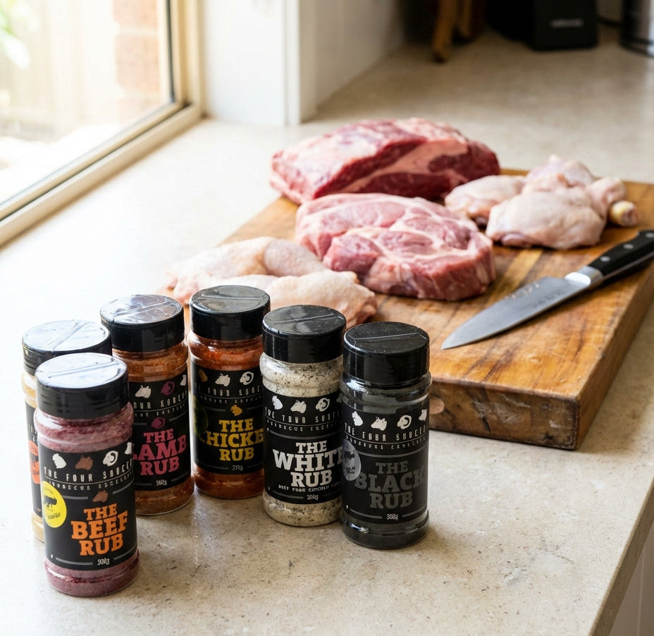 Six bottles of The Four Saucemen's seasoning rub set arranged on a kitchen counter in front of a wooden cutting board with raw meat and a knife.