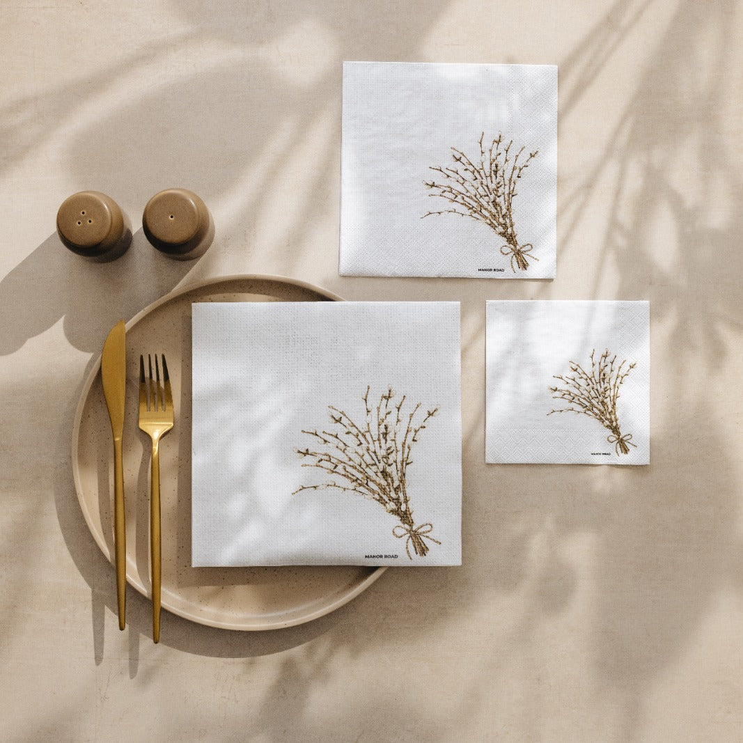 A styled flat-lay of white paper table linens with a printed botanical branch design, arranged on a beige surface with gold-toned cutlery, a plate, and small brown containers under soft leaf shadows.