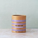 Olsson's Sea Salt Rub 'It's So Rosemary' in a kraft paper cylindrical container with lavender purple bands, set on a light marble surface against a textured sage green background.