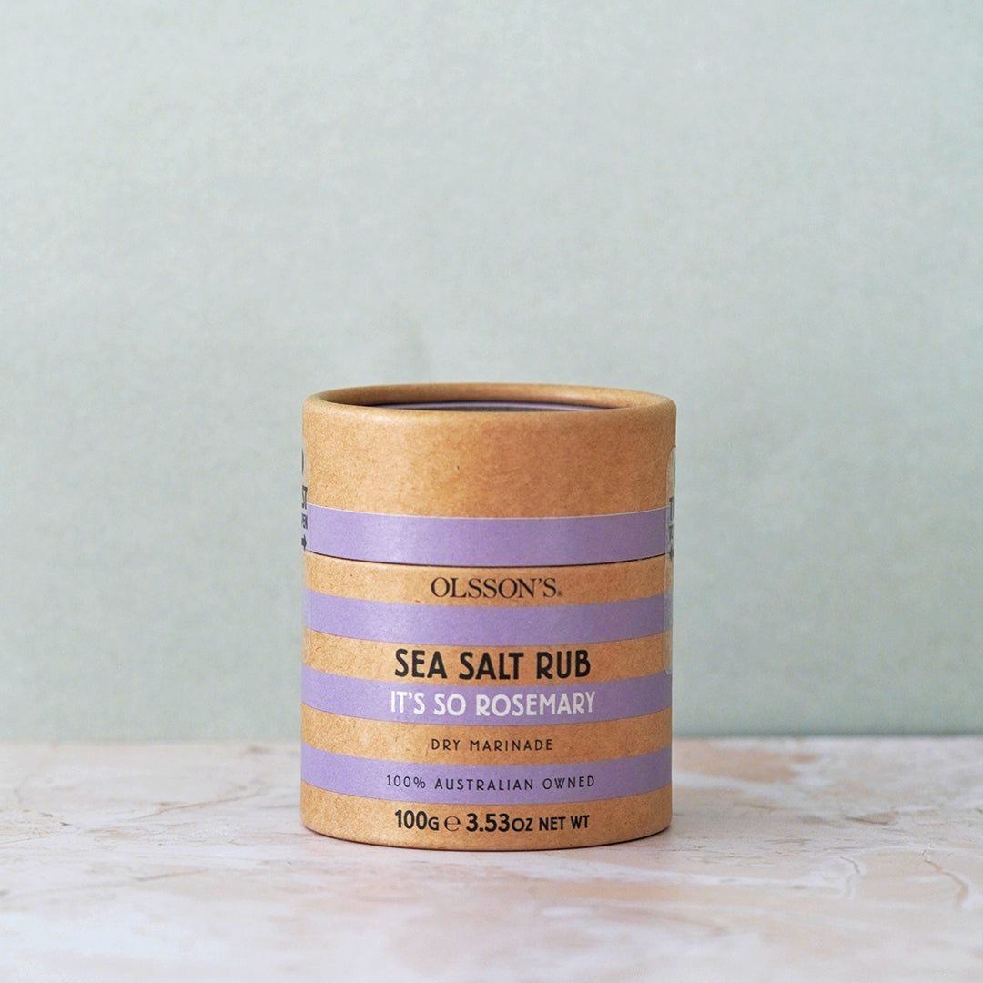 Olsson's Sea Salt Rub 'It's So Rosemary' in a kraft paper cylindrical container with lavender purple bands, set on a light marble surface against a textured sage green background.