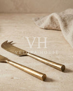 A gold-toned metal cheese knife and matching fork with textured handles, styled on a beige stone surface with a neutral cloth and plate in the background.