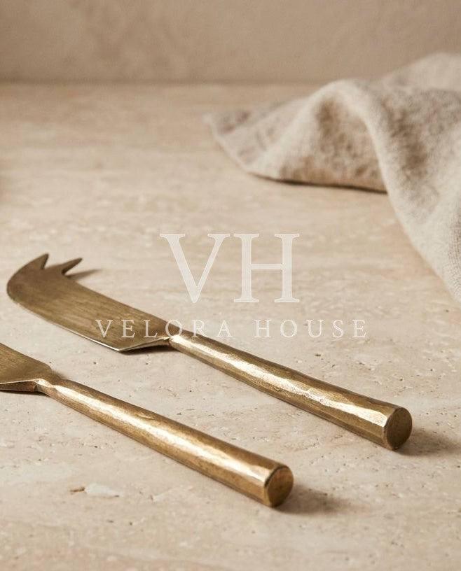 A gold-toned metal cheese knife and matching fork with textured handles, styled on a beige stone surface with a neutral cloth and plate in the background.